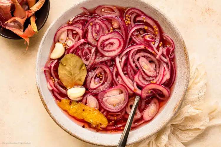 Photo of a quick pickled red onion with garlic, bay leaf, and lemon peel in a white bowl
