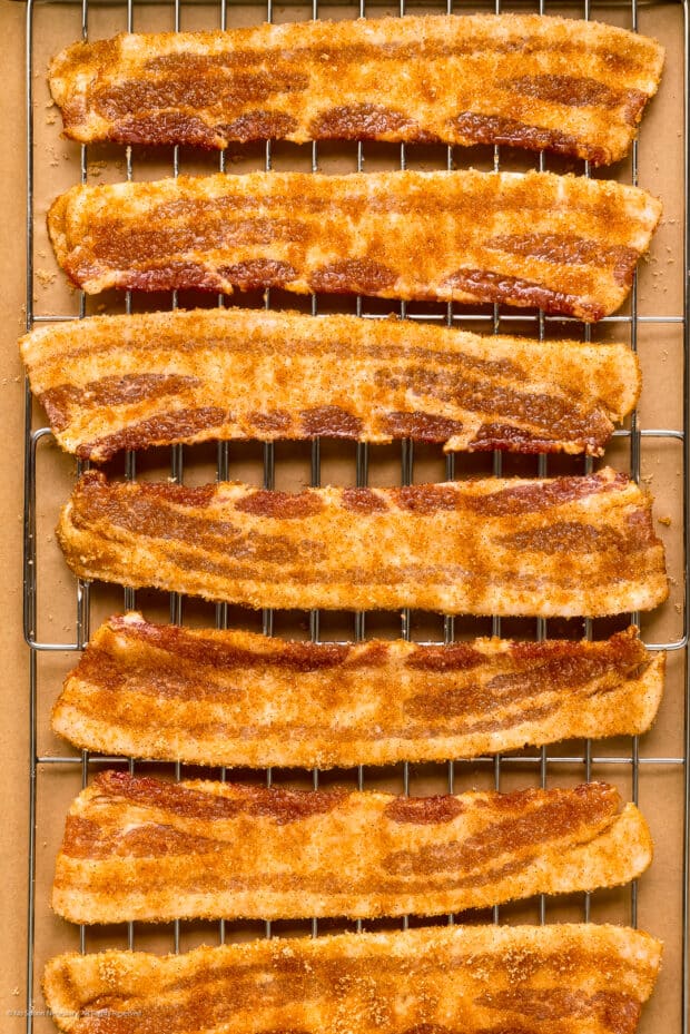 Photo of seven strips of raw bacon brushed with maple syrup and sprinkled with brown sugar on a wire rack.