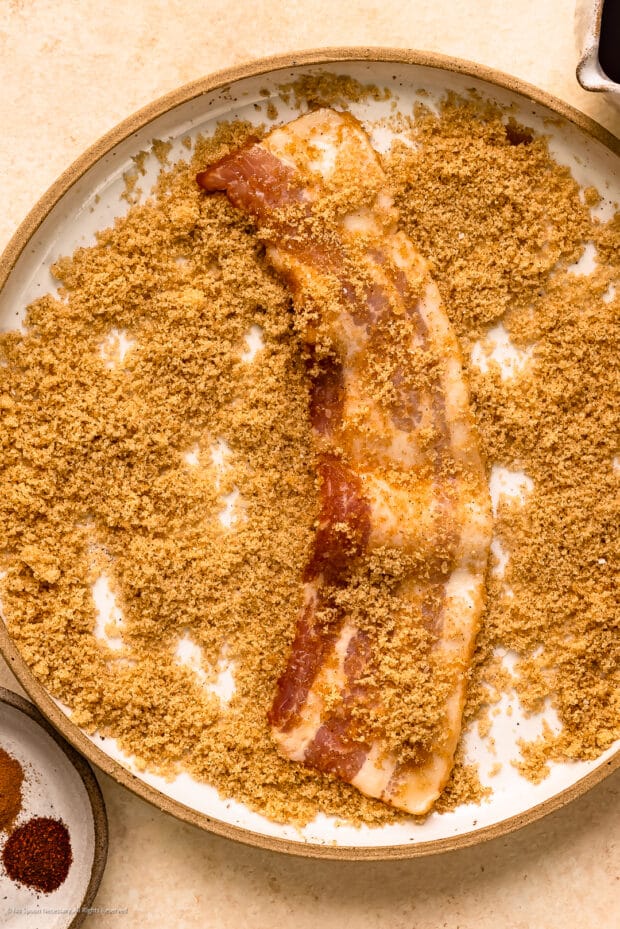 Overhead photo of raw strip of bacon in a bowl of brown sugar. 