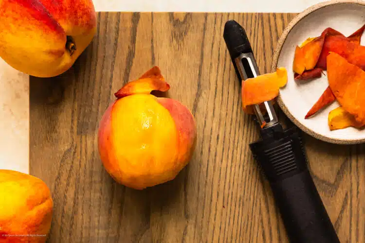 Overhead photo of a partially peeled peaches on a cutting board with a vegetable peeler next the to fruit.