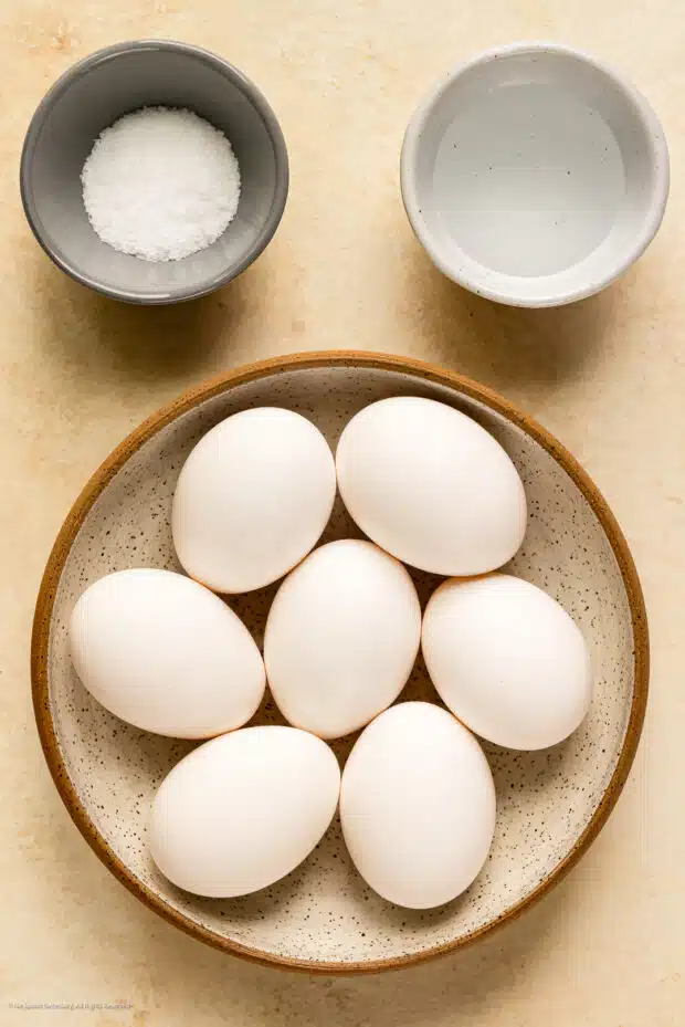 Overhead photo of six raw eggs, a ramekin of kosher salt, and a ramekin of white vinegar on a kitchen counter.