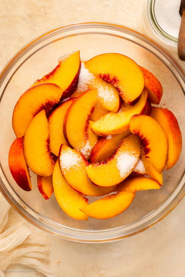 Overhead photo of fresh slices of peaches sprinkled with granulated sugar in a large mixing bowl.
