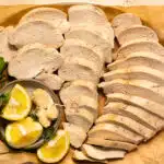 Overhead photo of two pounds of poached chicken breasts thinly sliced and arranged on a serving platter with lemon wedges and fresh herbs.