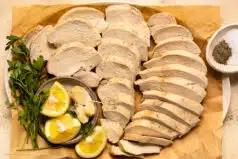 Overhead photo of two pounds of poached chicken breasts thinly sliced and arranged on a serving platter with lemon wedges and fresh herbs.