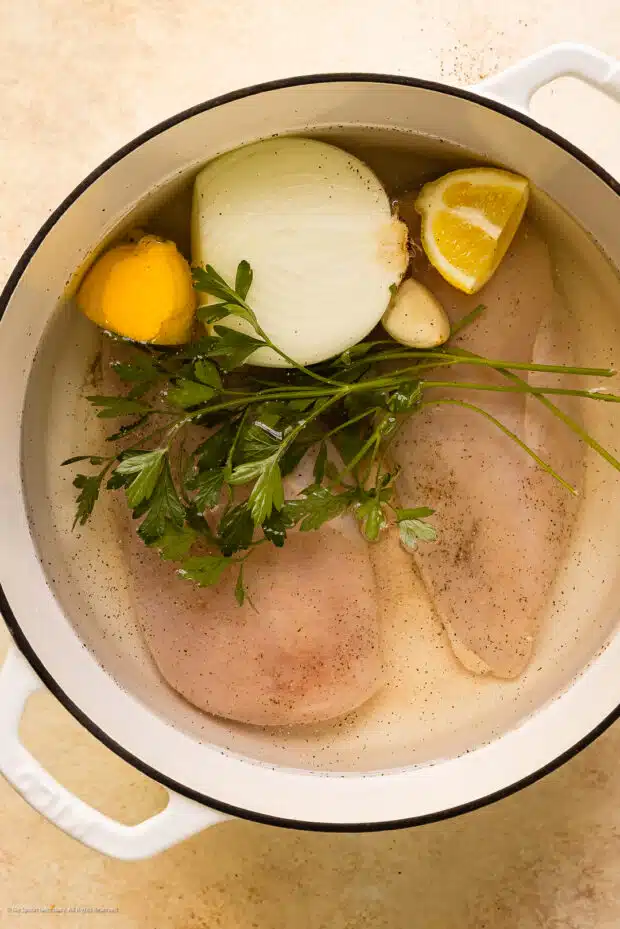 Action photo of showing two chicken breasts poaching in water with lemon wedges, garlic cloves, half an onion, and a handful of fresh parsley.