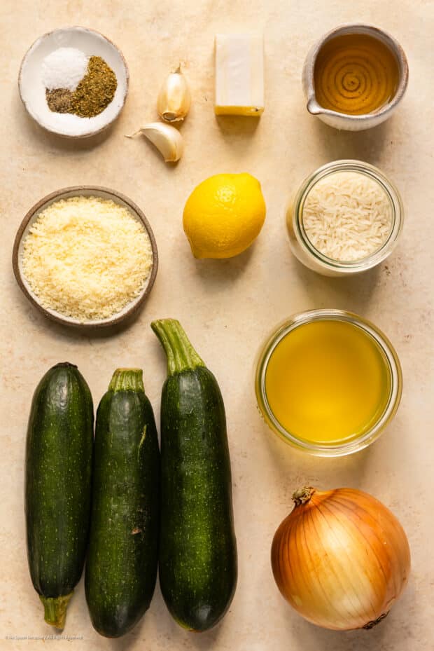 Overhead photo of 3 zucchini, a yellow onion, broth, white rice, parmesan cheese, butter, and seasonings on a kitchen counter.