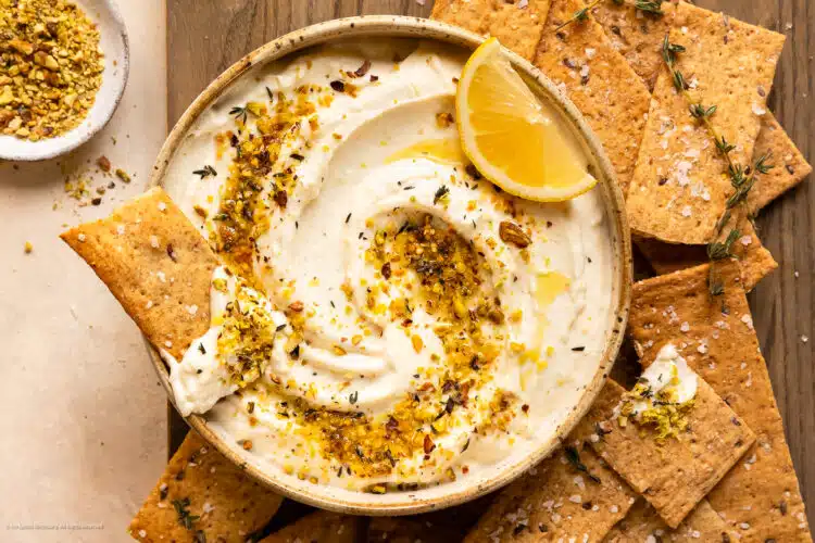 Overhead photo of a bowl of whipped ricotta topped with lemon zest and fresh herbs with crackers on the side of the bowl.