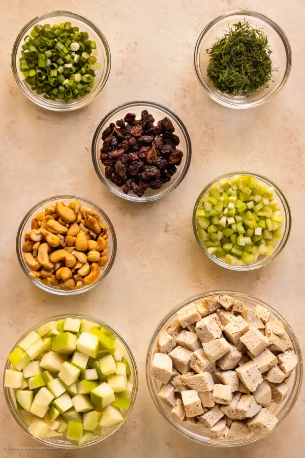 Overhead photo of chopped cooked chicken breasts, apples, celery, cashews, raisins, and fresh herbs neatly arranged in bowls on a kitchen counter.