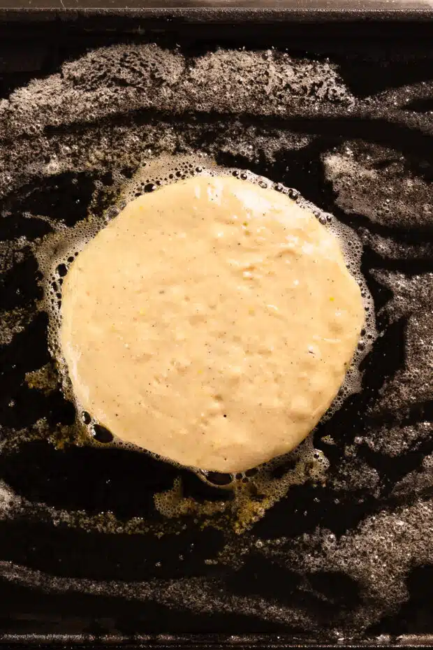 Action photo of a circle of raw pancake batter cooking on a hot buttered pan.