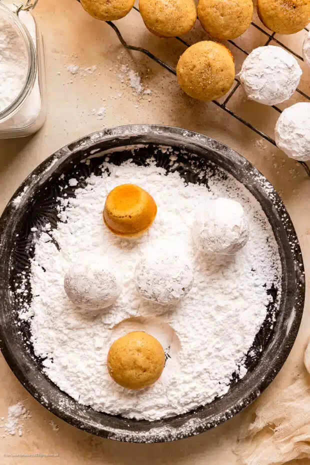 Action photo of baked donut holes being roll in powdered sugar.