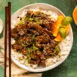 Overhead photo of crispy orange beef over white rice with orange wedges in a white bowl.