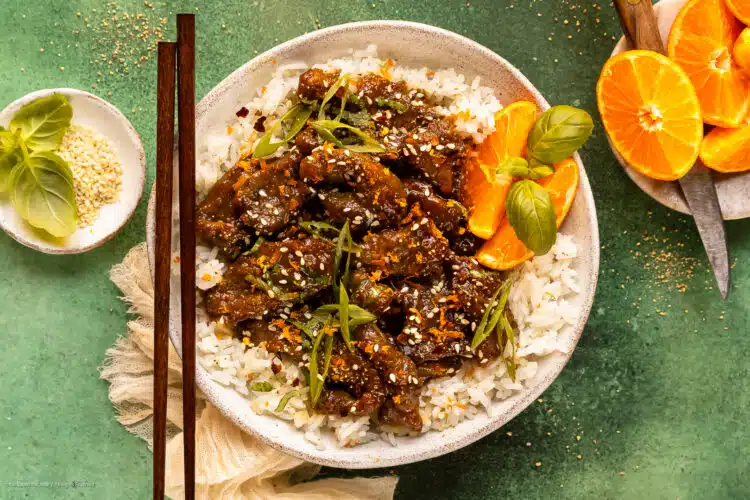 Overhead photo of crispy orange beef over white rice with orange wedges in a white bowl.