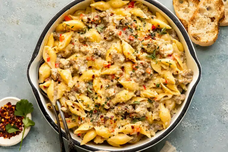 Overhead, landscape photo of creamy sausage pasta garnished with fresh parsley in a large white skillet with slices of crusty bread next to the pan.