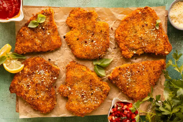 Overhead photo of six parmesan crusted chicken cutlets garnished with fresh basil on a serving board.