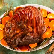 Overhead photo of an orange glazed ham on a white serving platter with fresh oranges, pomegranate arils, rosemary, and thyme.