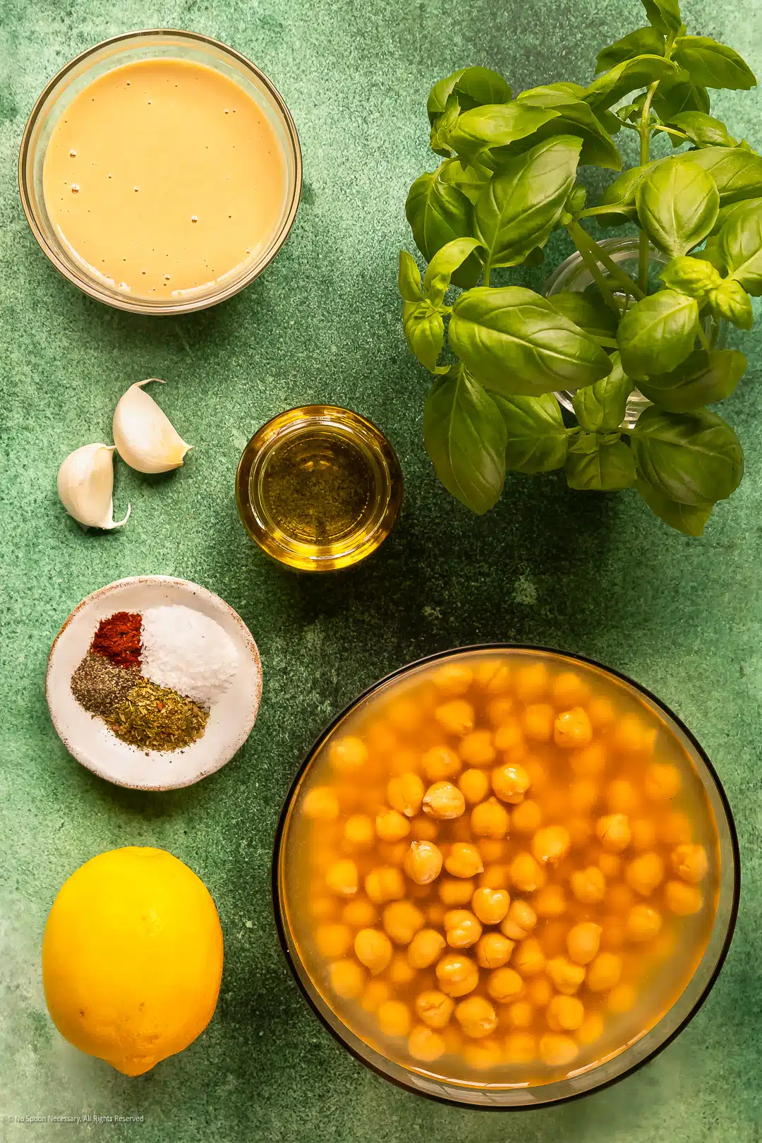 Overhead photo of a fresh basil plant, canned chickpeas, tahini, garlic cloves, and spices neatly arranged on a kitchen counter.